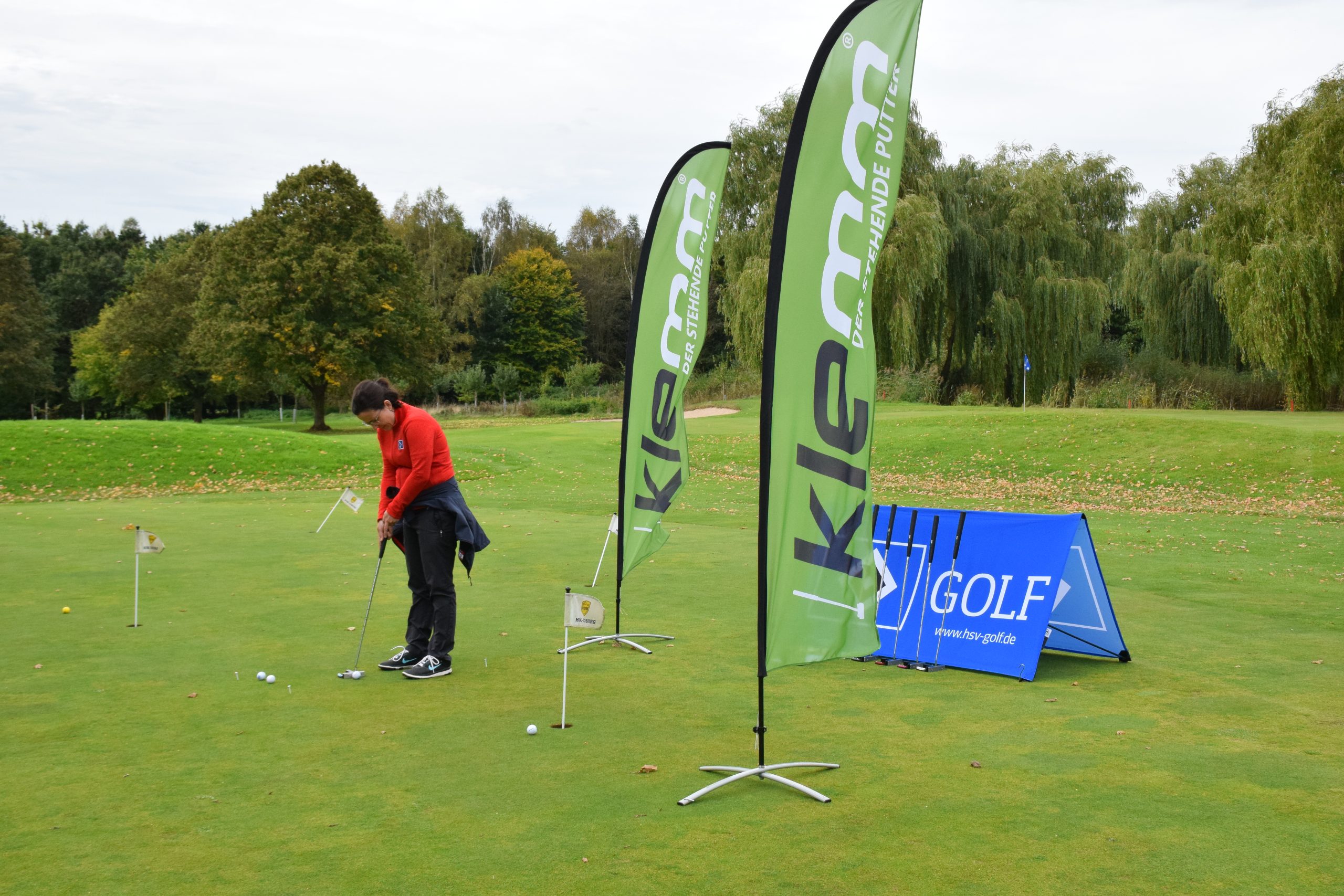 Golferin hat einen Putter von Klemm - Der stehende Putter in der Hand und puttet. Rechts daneben stehen zwei Beachflags mit Logo der Firma darauf.